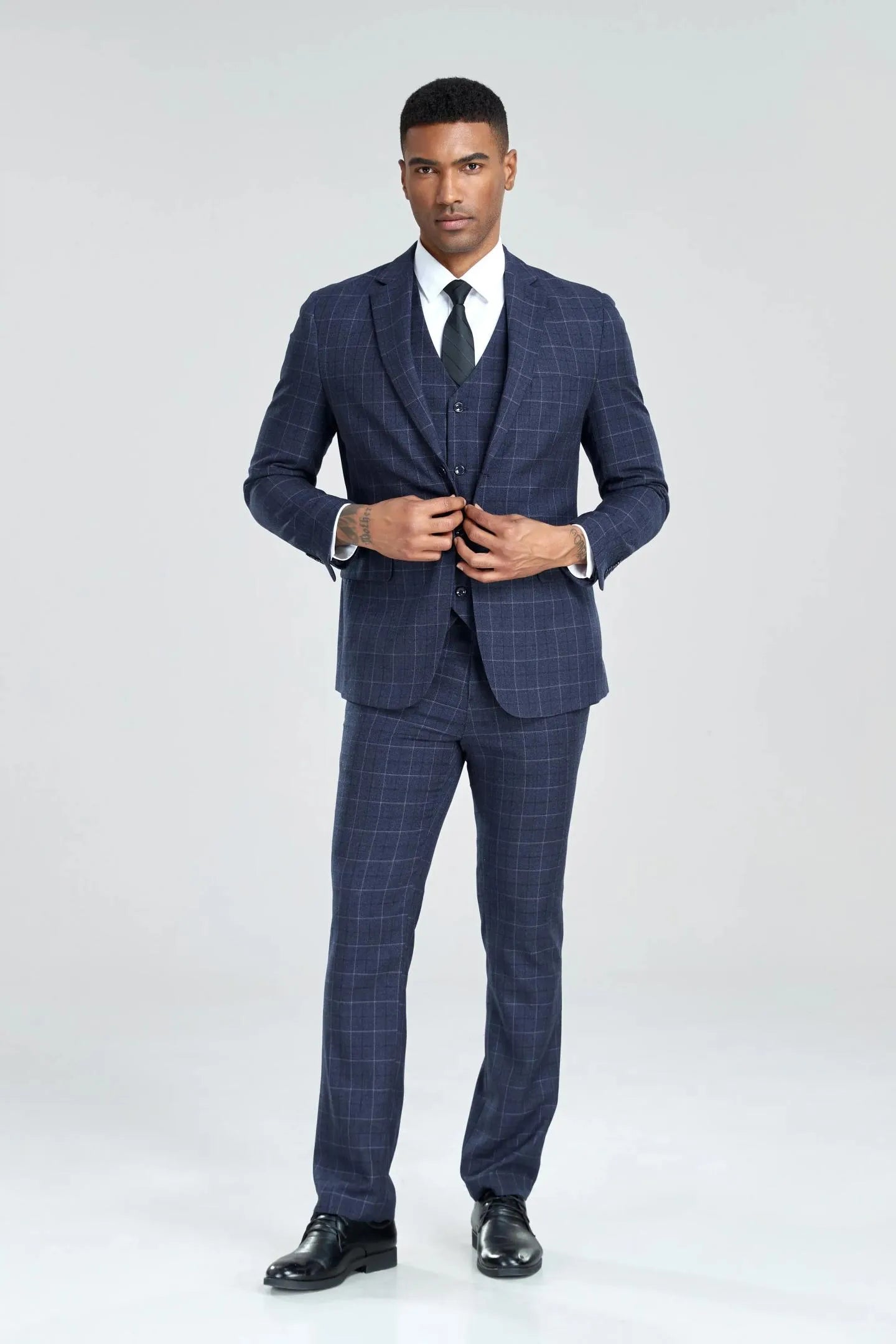 model wearing navy plaid 3-piece suit, adjusting vest buttons, black tie complementing the formal business attire.