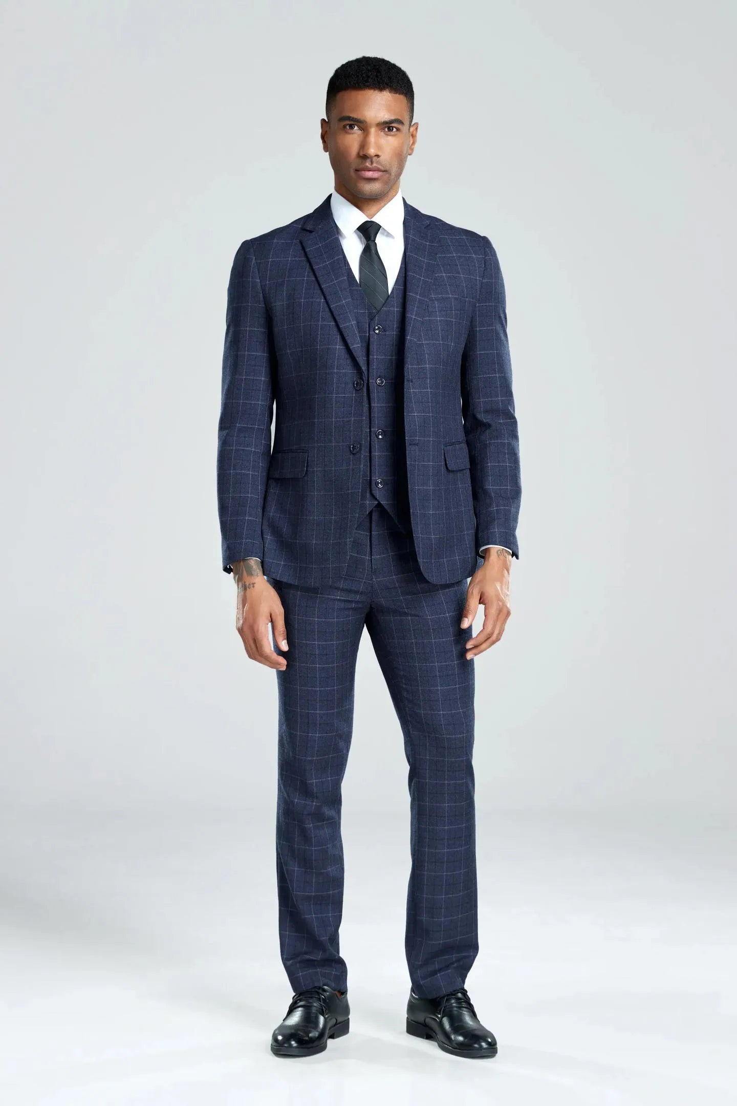 full view of model in navy plaid 3-piece suit, buttoning jacket, paired with black tie for a formal look.