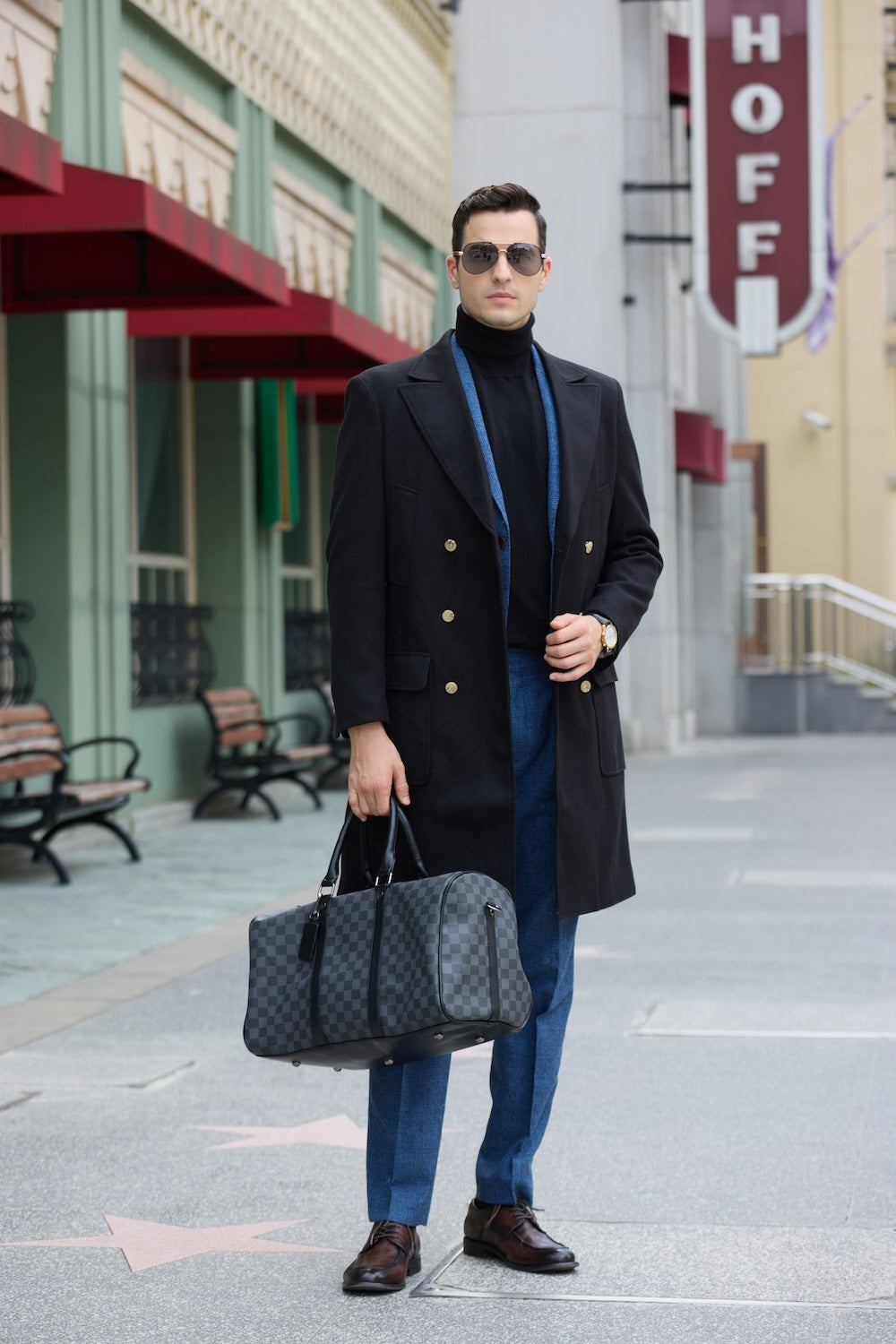 Full body shot of man in black long coat holding travel bag in urban outdoor scene