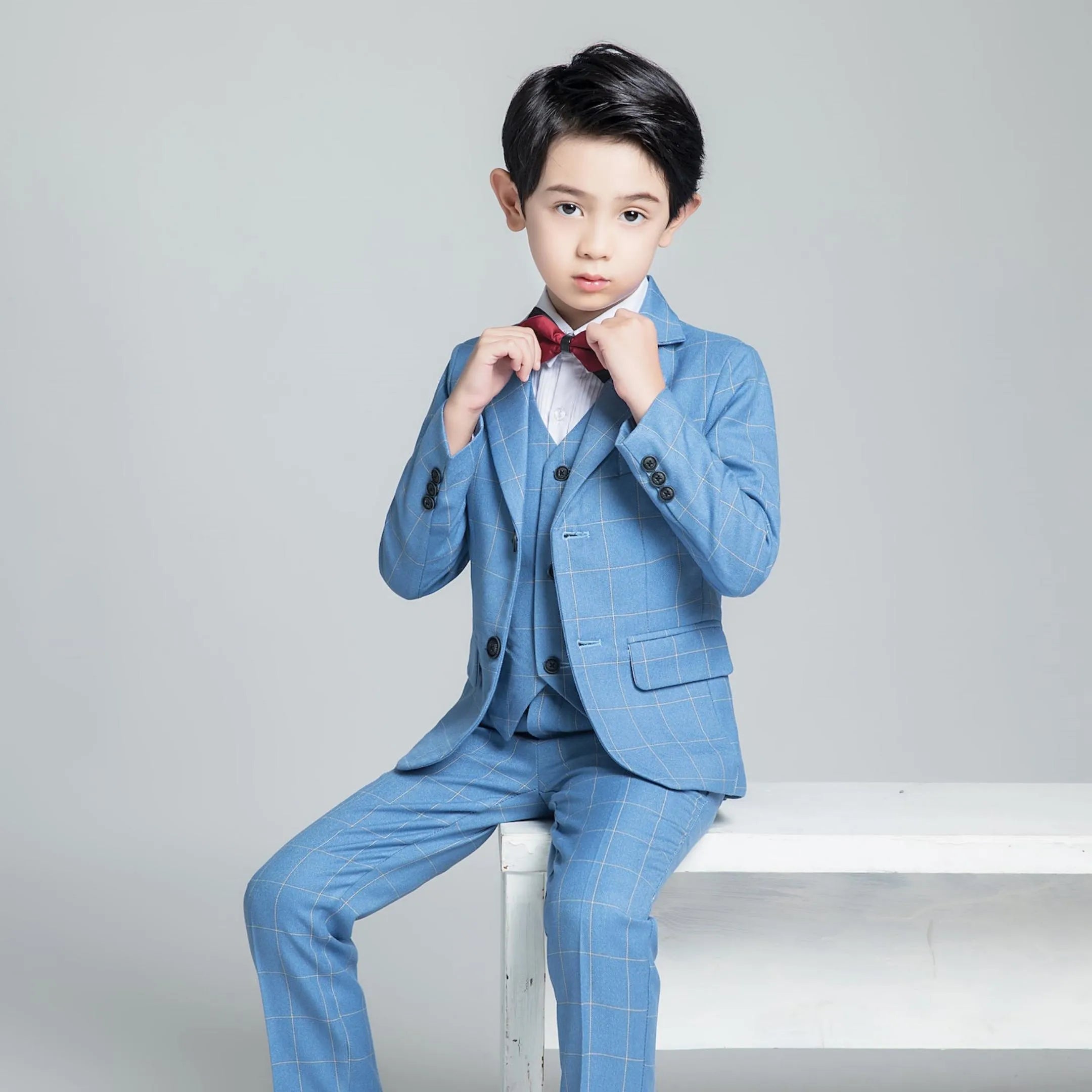 Sitting pose of boy in elegant 5-piece suit adjusting red bow tie