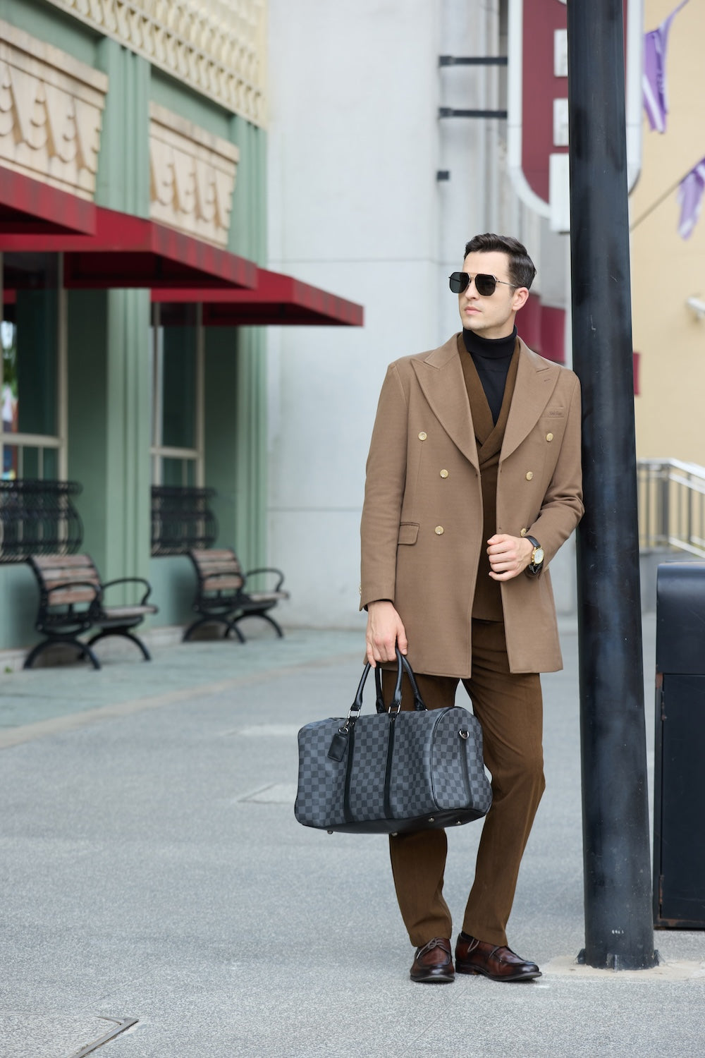 Men's coffee wool overcoat - side view of double breasted long winter business coat, holding checked duffle bag by urban street lamp