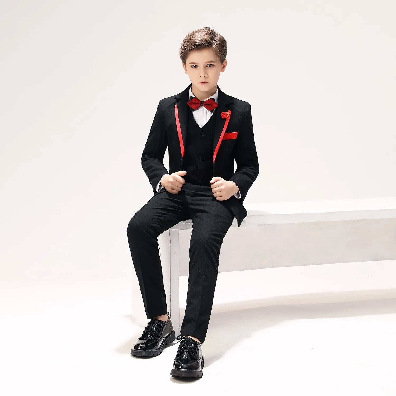 Boys' tuxedo 5-piece black formal set with red accents, bow tie and vest, seated pose with glossy dress shoes