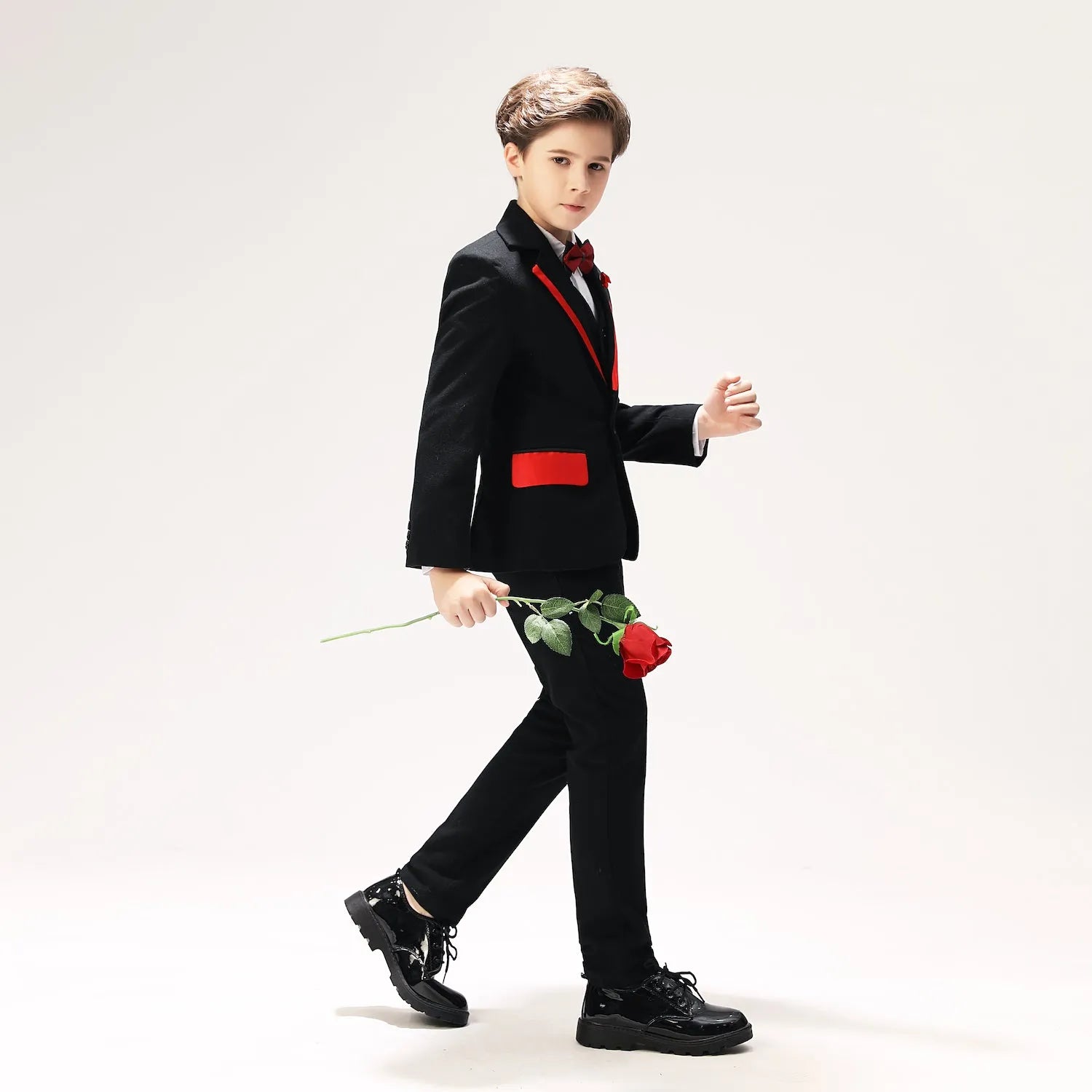 Boys' prom suit with red trim and bow tie, black formal outfit styled with rose, walking pose in glossy shoes