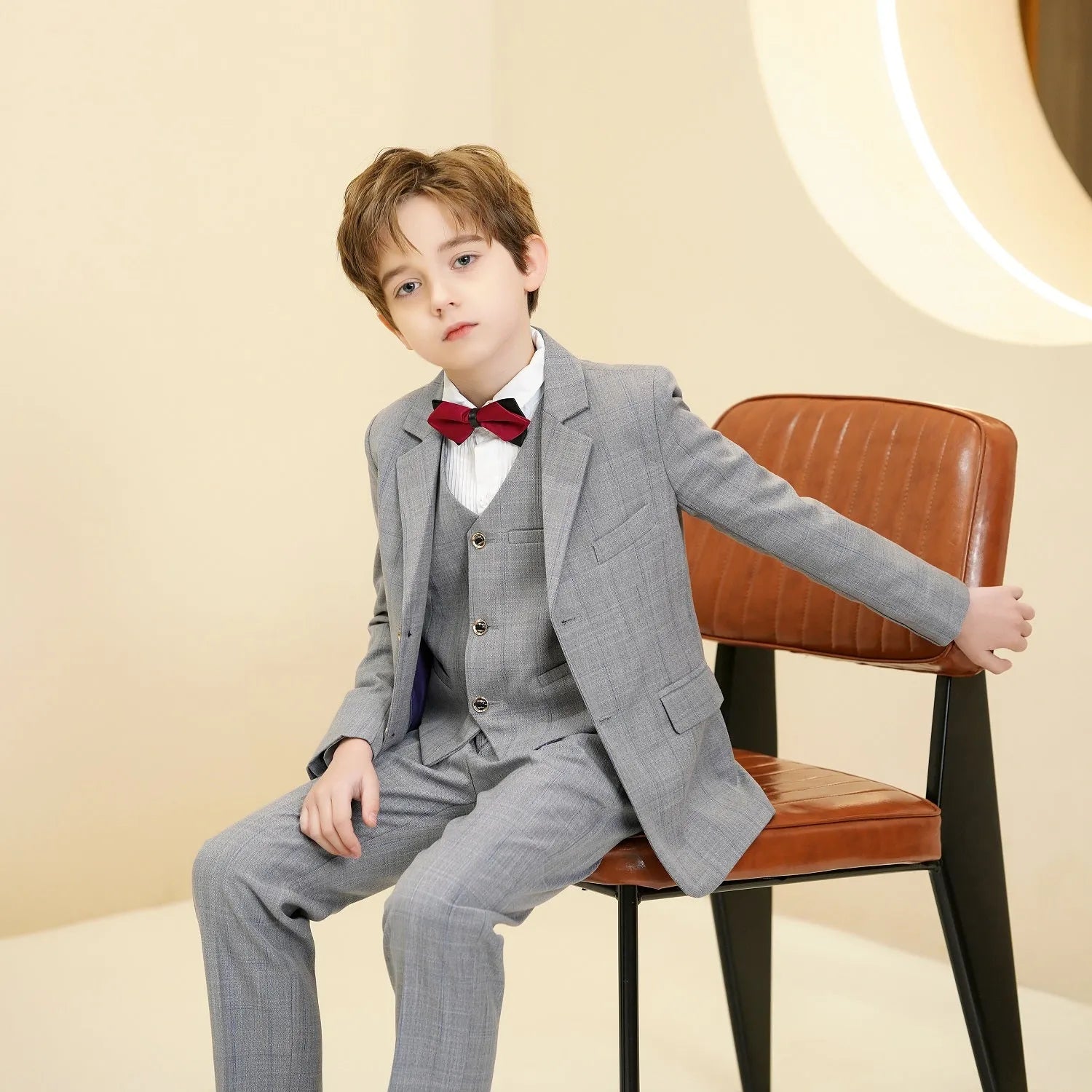 Young boy seated in light grey 5 piece plaid suit with vest and bow tie for formal events.