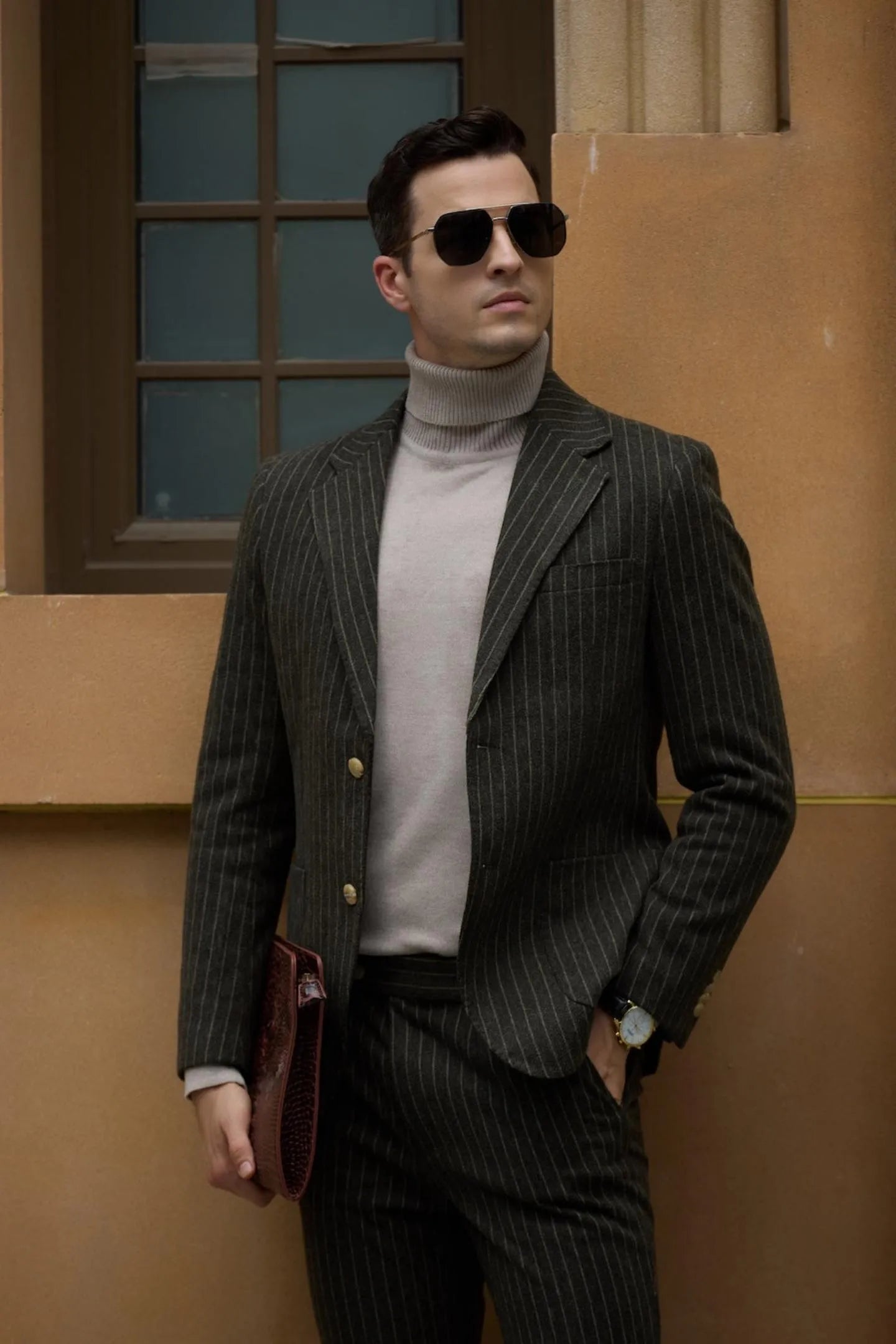 Close-up of a man wearing a men's green herringbone pinstripe 2-piece suit with a beige turtleneck, holding sunglasses and showcasing the fine suit details.