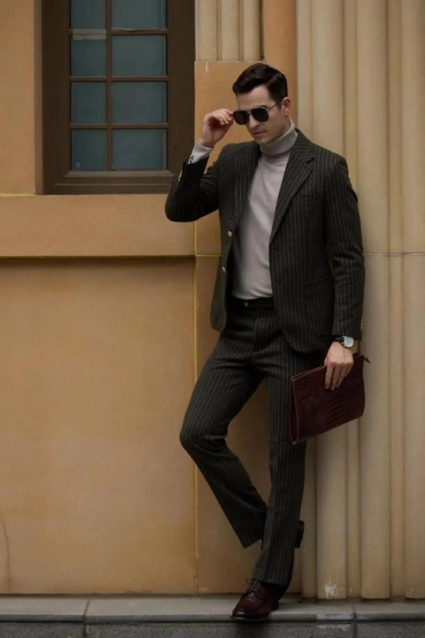 Man wearing a men's green herringbone pinstripe 2-piece suit, standing in a relaxed pose with sunglasses, carrying a briefcase.
