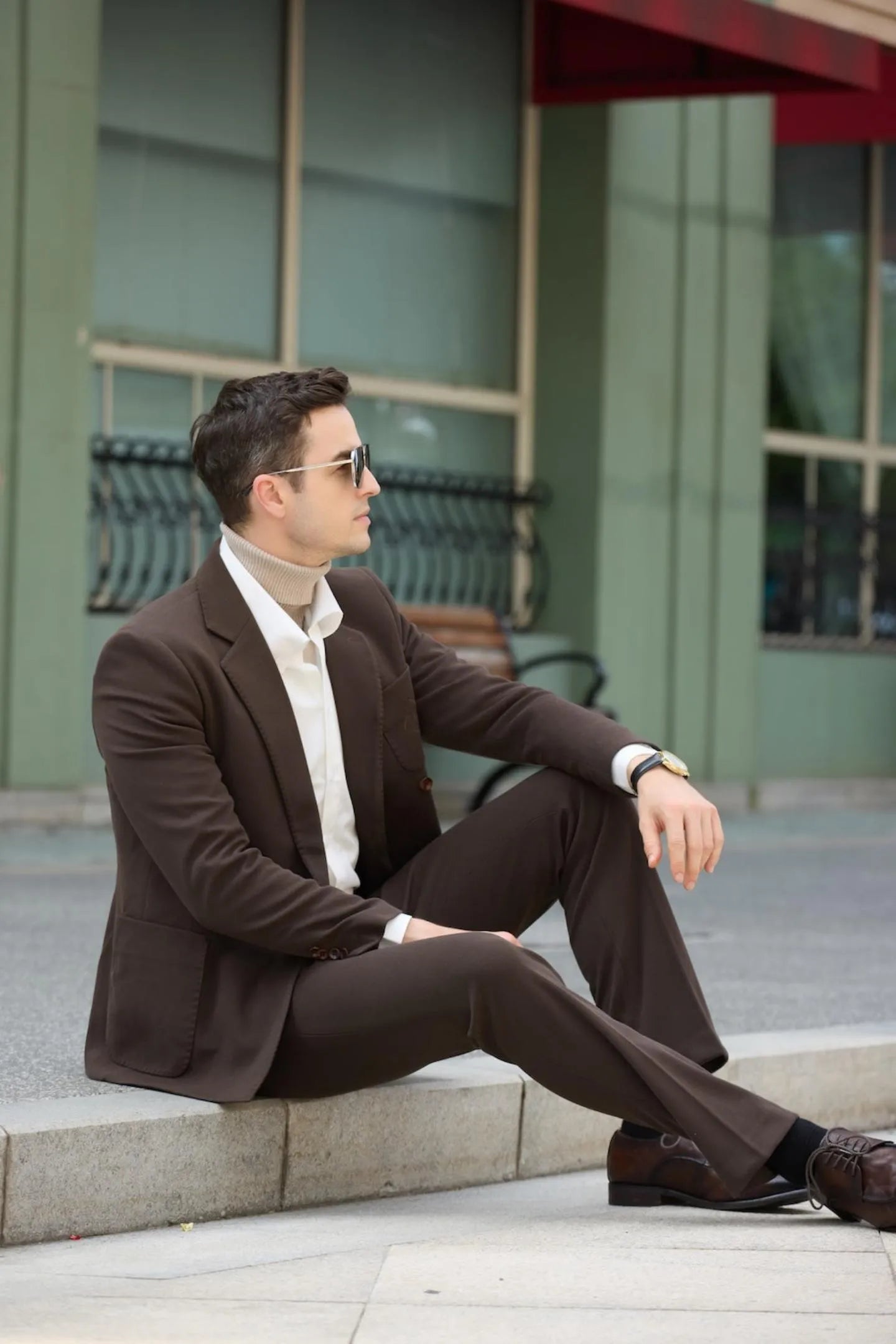 Model sitting while wearing coffee herringbone tweed 2 piece men's suit, featuring tailored trousers and blazer