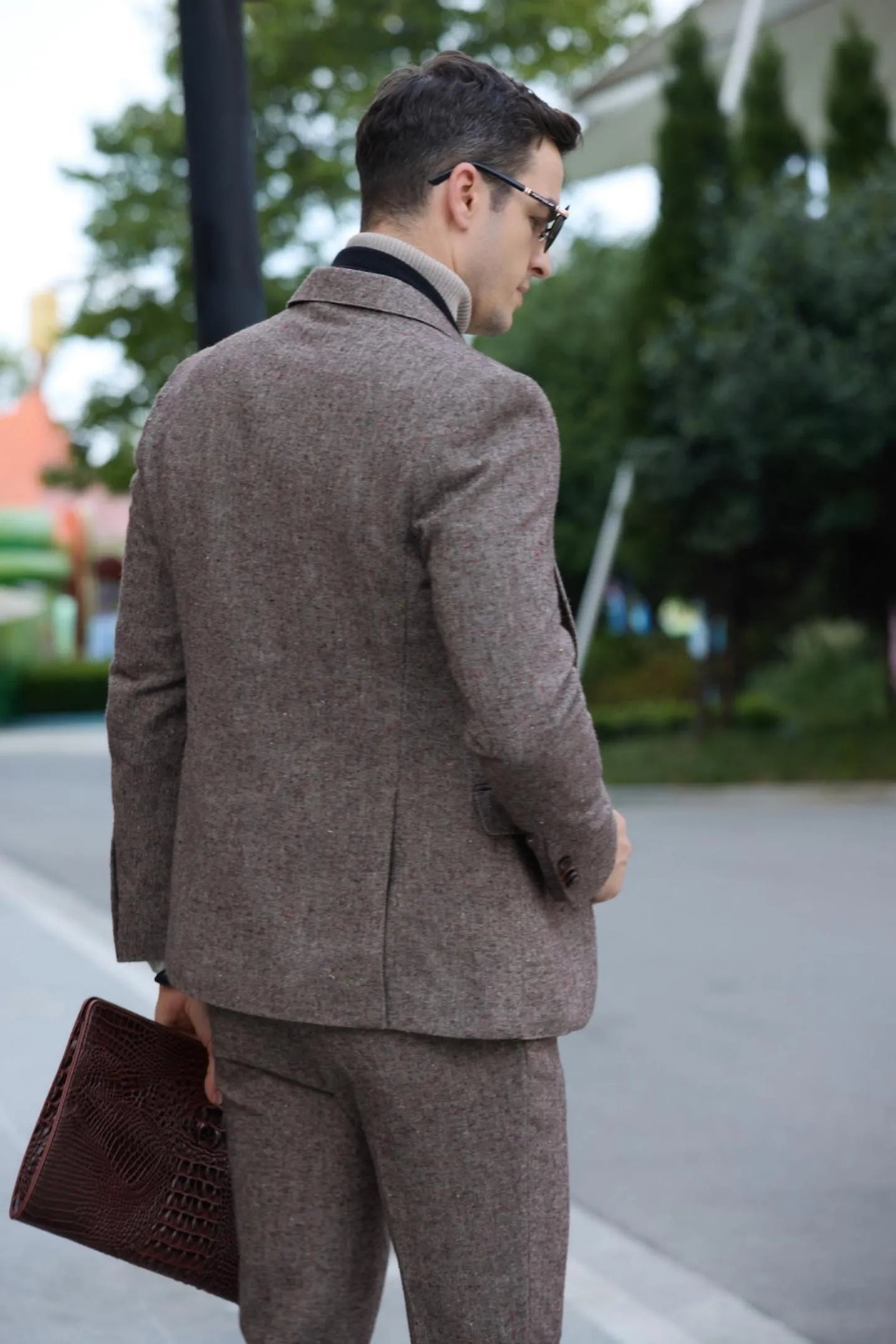 back view of men's brown tweed herringbone 2-piece suit with black scarf and leather clutch, standing casually outdoors.