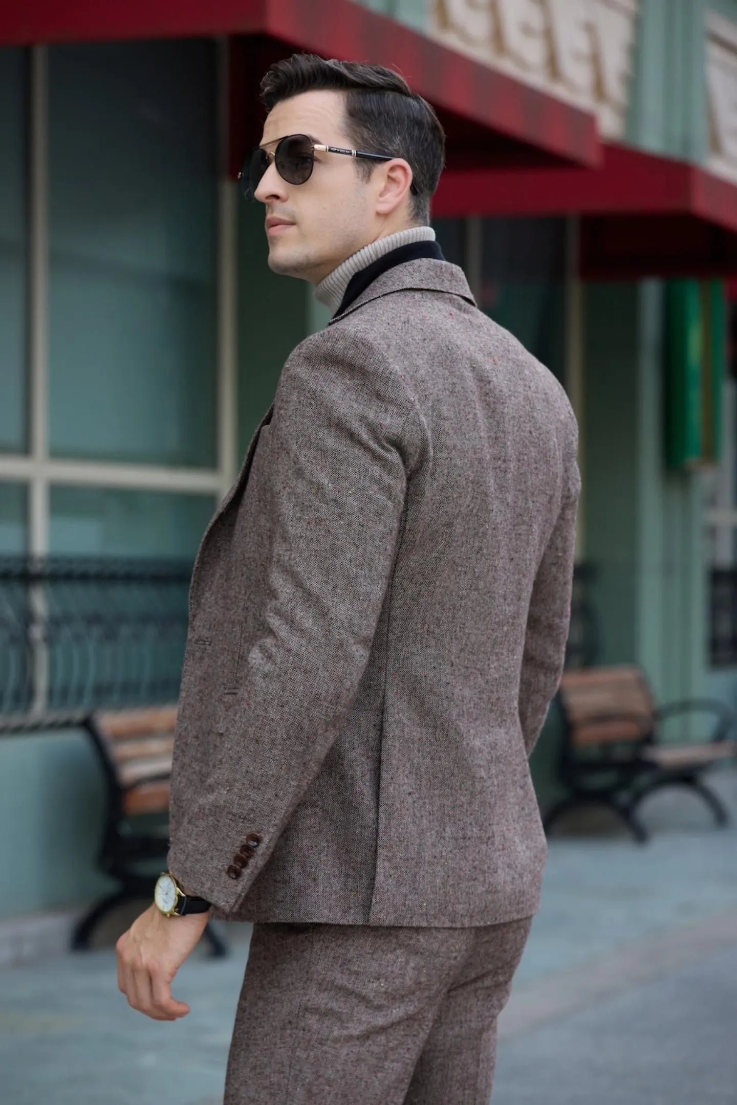 side view of men's brown tweed herringbone 2-piece suit with black scarf and leather clutch, walking in the city.