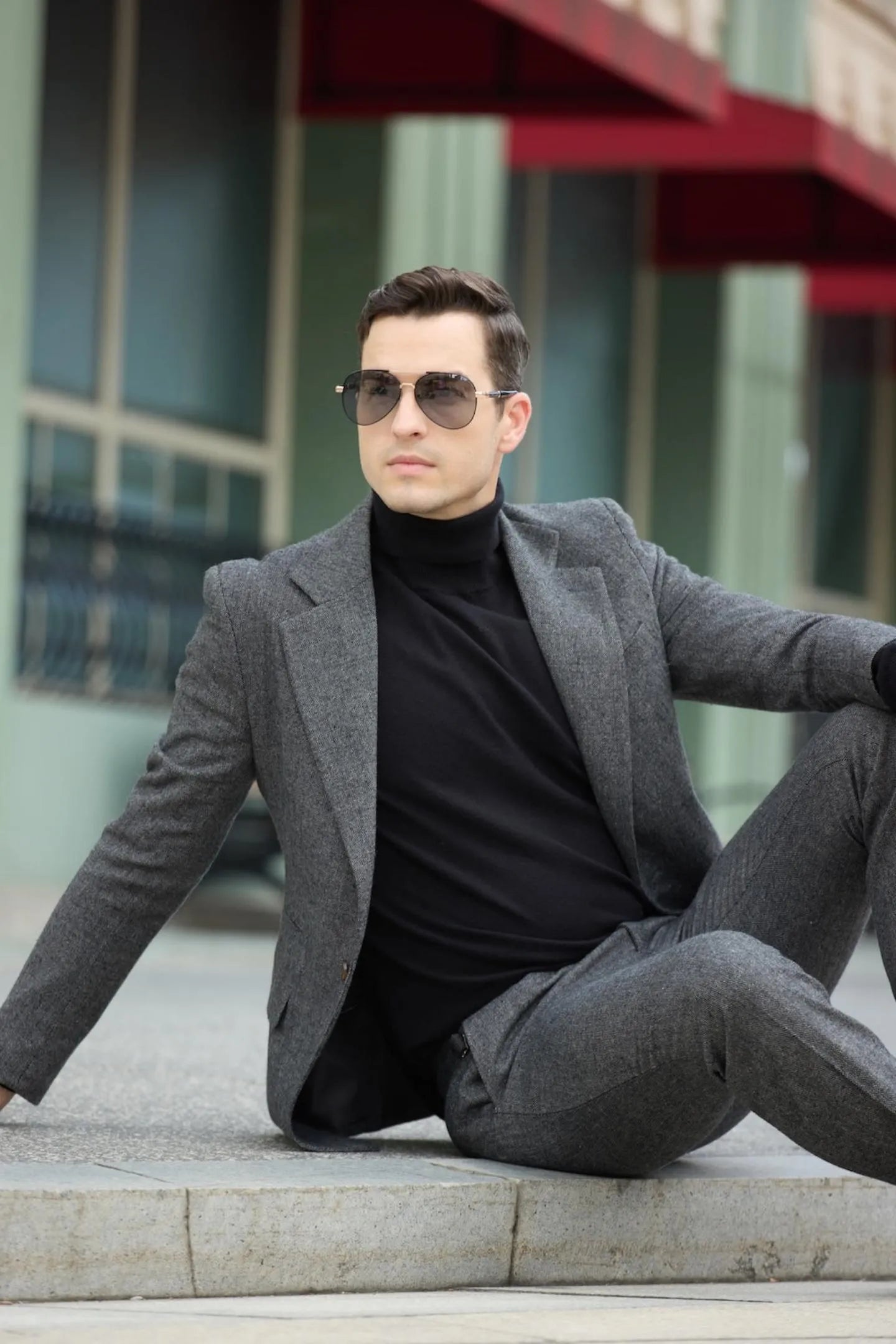 model seated in men's dark grey tweed herringbone 2 piece suit, black turtleneck, and sunglasses.
