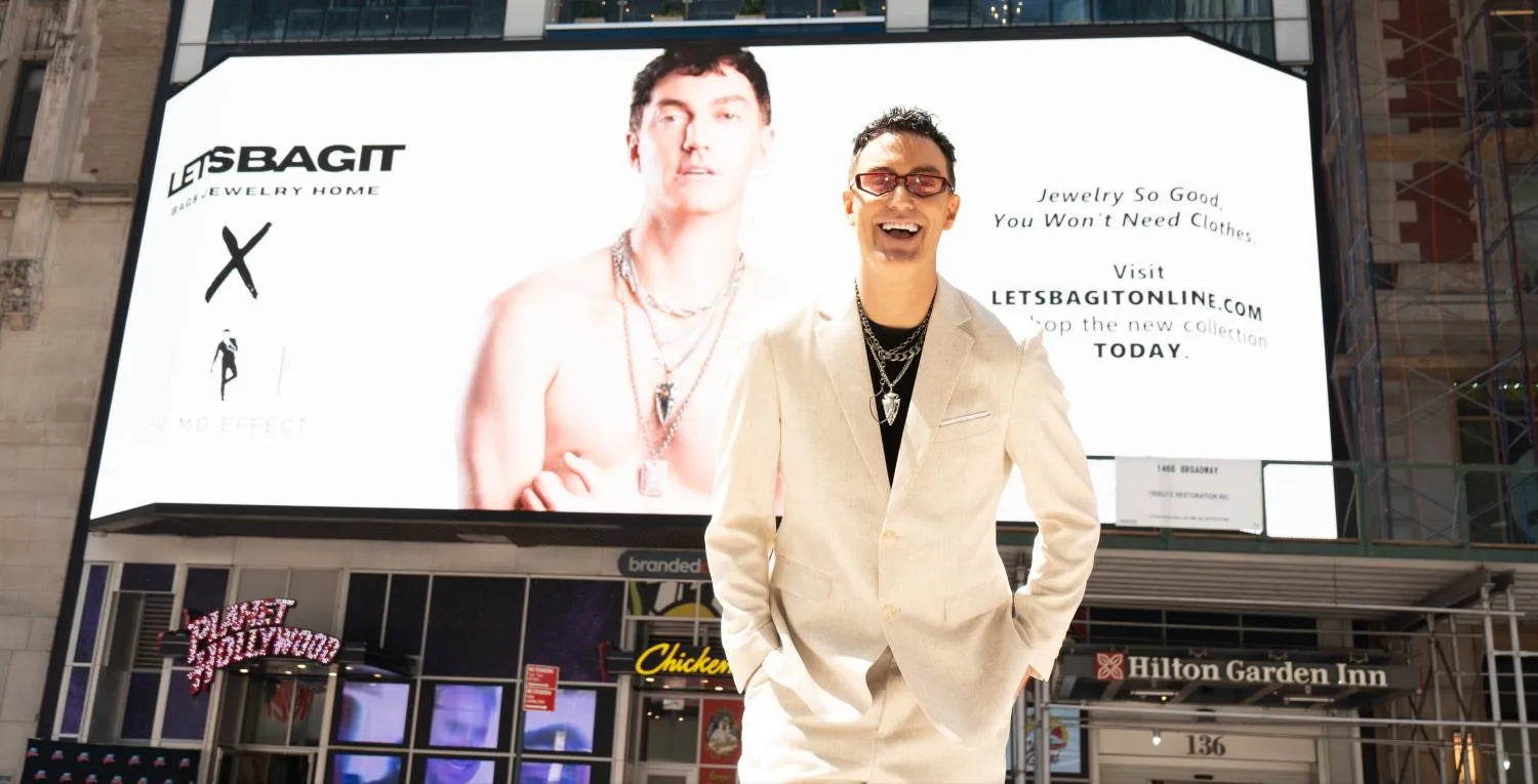 Stylish man in a light-beige Wehilion suit, layered necklaces, and sunglasses smiling in front of a giant city billboard.