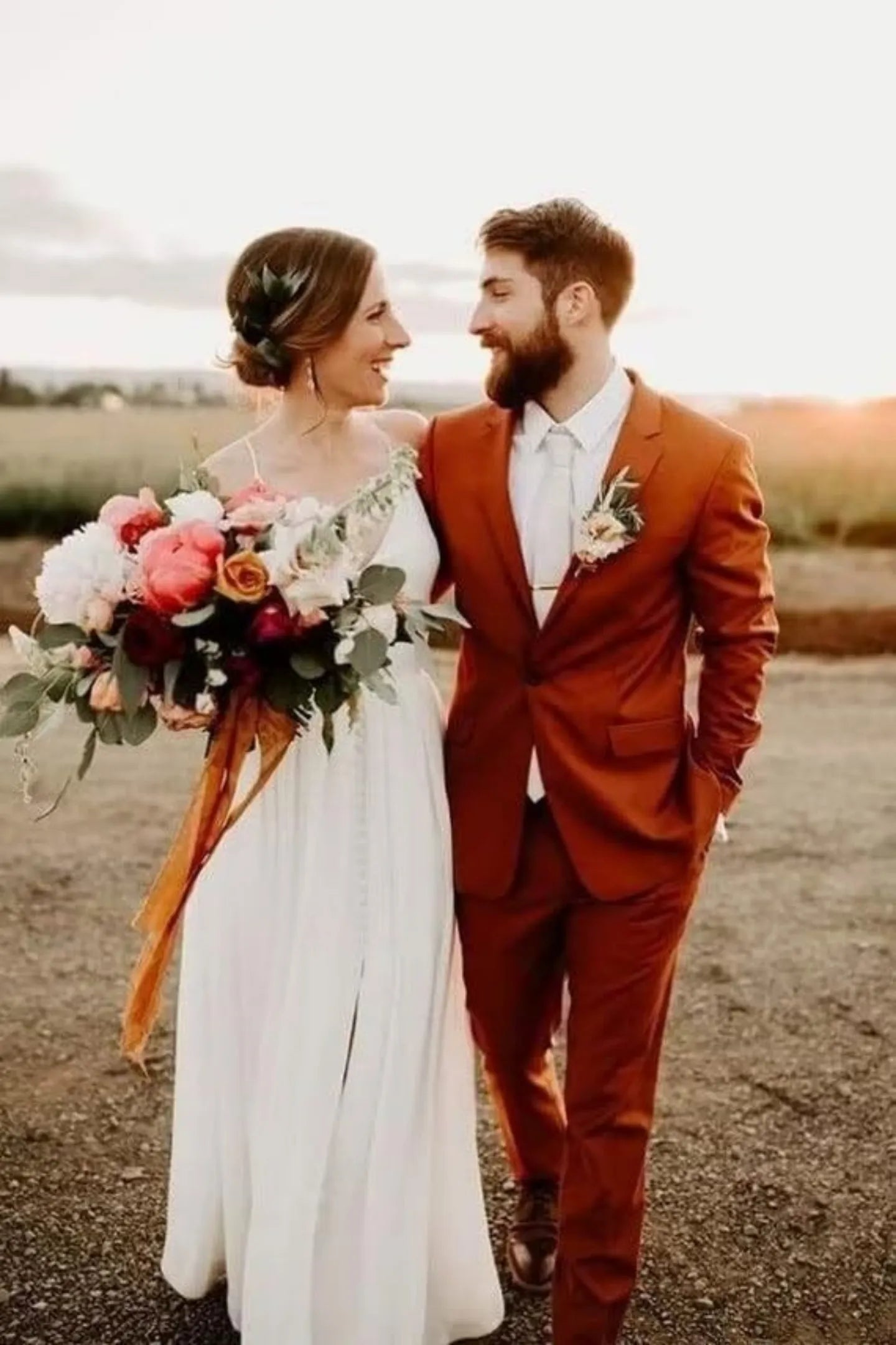 groom in burnt orange slim tuxedo suit holding bride with floral bouquet at sunset