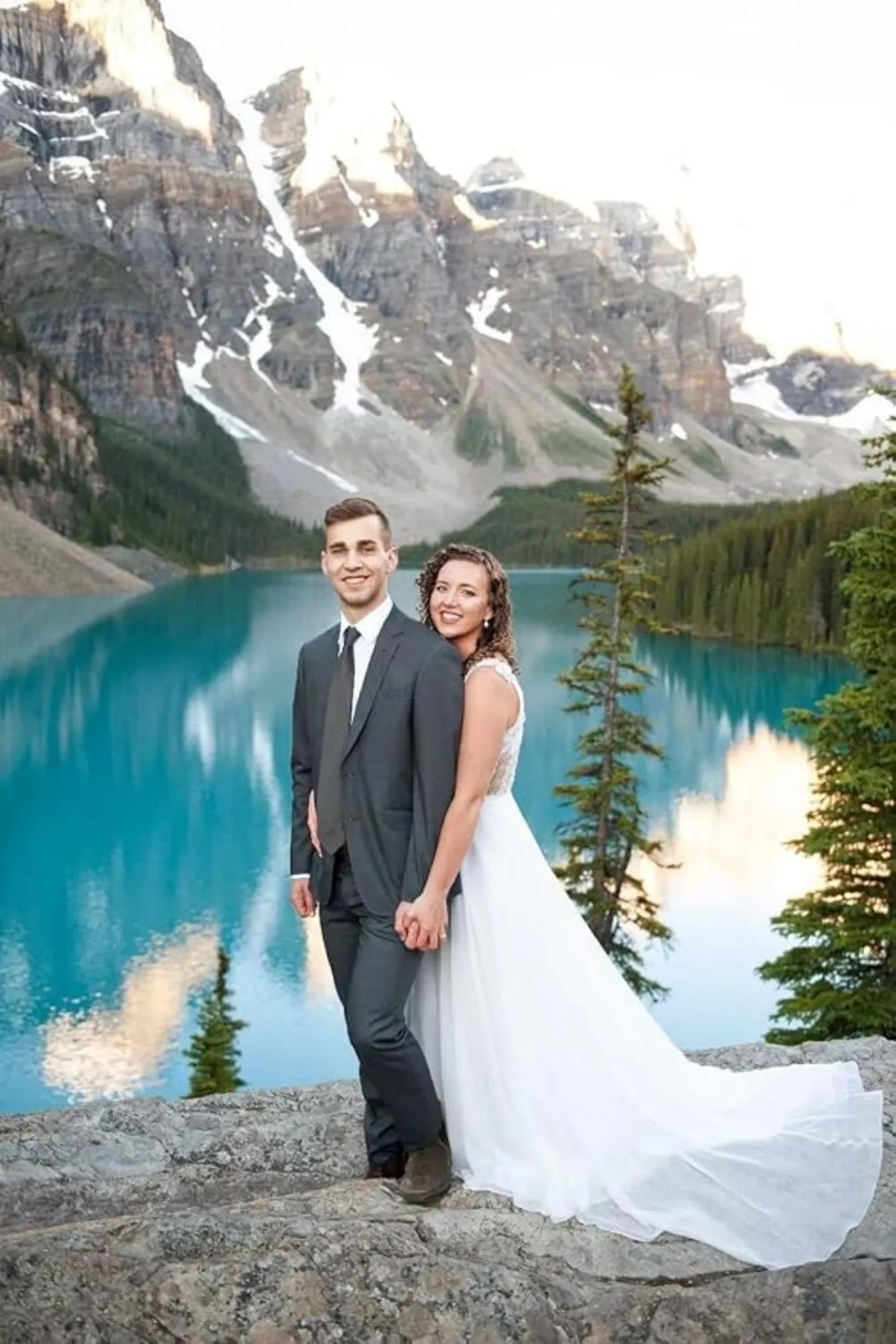 groom in grey 3 piece wedding suit with bride by alpine lake and mountain backdrop