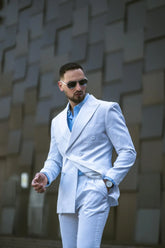 Confident man in a light-blue double-breasted suit and aviator sunglasses walking past a modern textured wall.