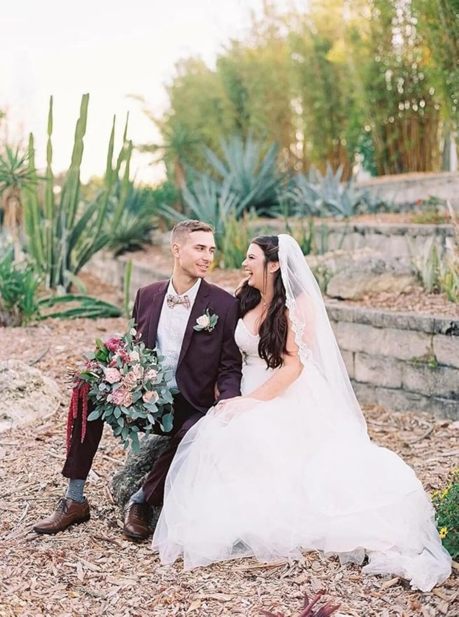 modern groom in dark purple 3 piece suit sitting with bride in romantic garden wedding