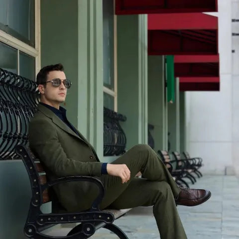 Stylish man in an olive-green three-piece suit sitting on an outdoor bench, showcasing modern men’s formalwear.