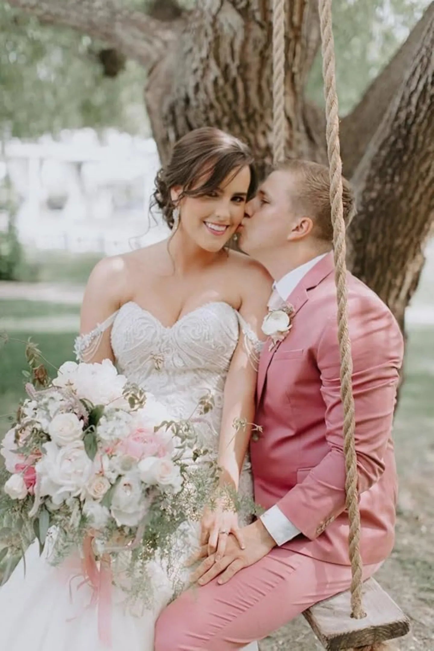 groom in pastel pink wedding suit sitting on swing with bride holding romantic floral bouquet