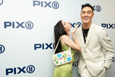 Smiling man in a light beige suit and black tee stands beside a woman in a lime green dress with a floral beaded bag, posing in front of a PIX11 logo backdrop.