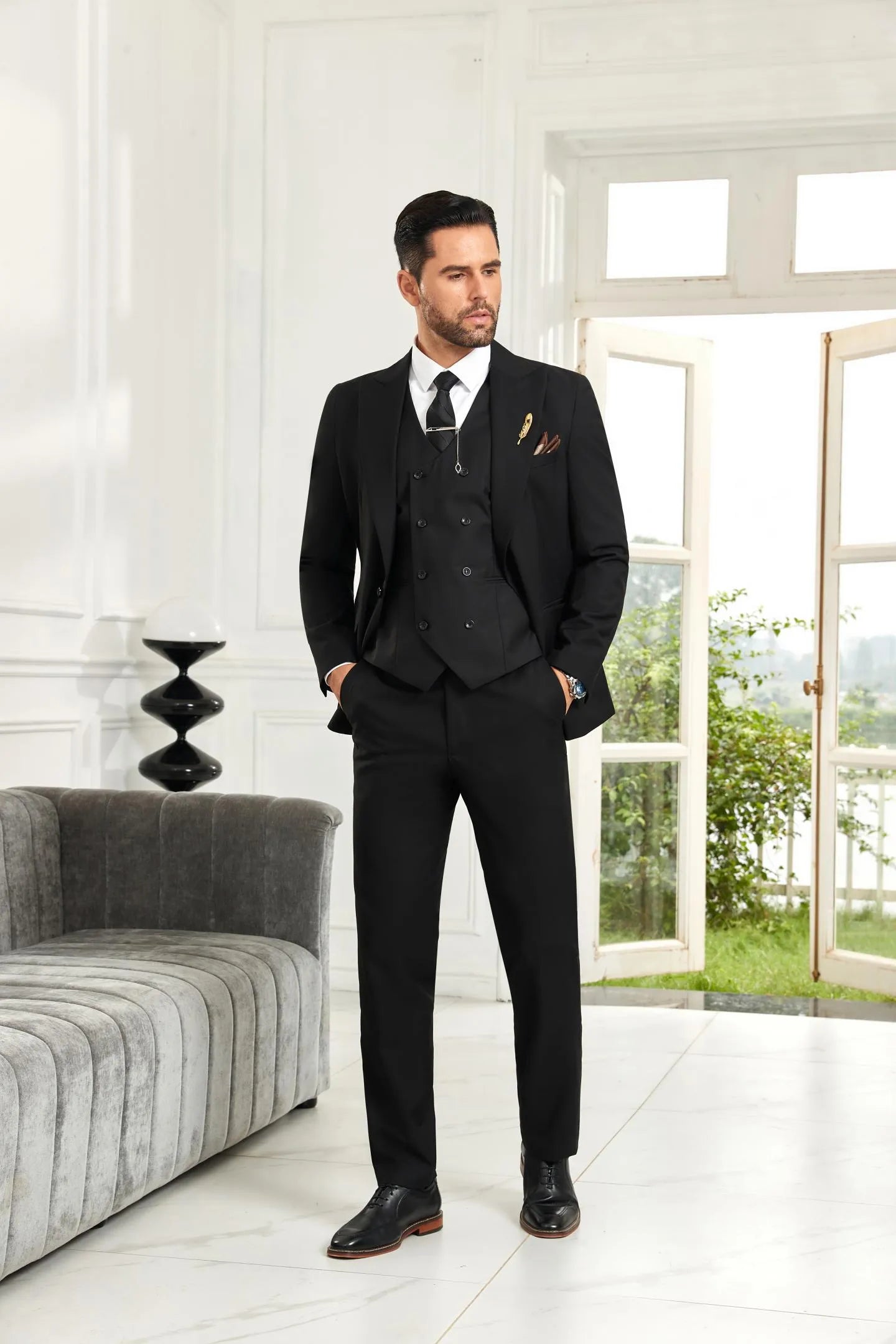 Black modern 3 piece men's suit with slim fit blazer and vest, model posing with hands in pockets indoors.