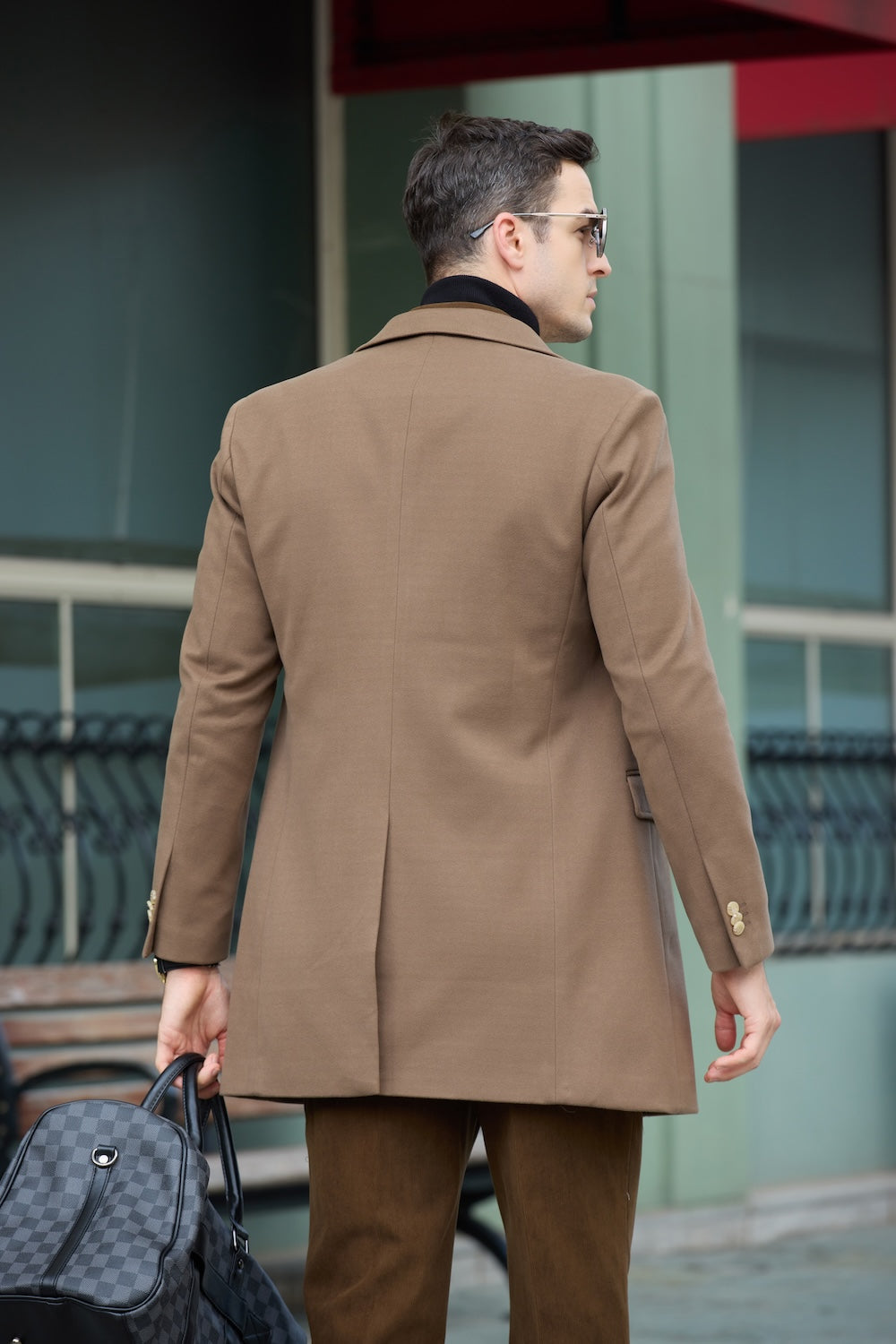 Men's coffee wool overcoat - back view of double breasted long winter business coat with center vent, holding checked bag in urban building