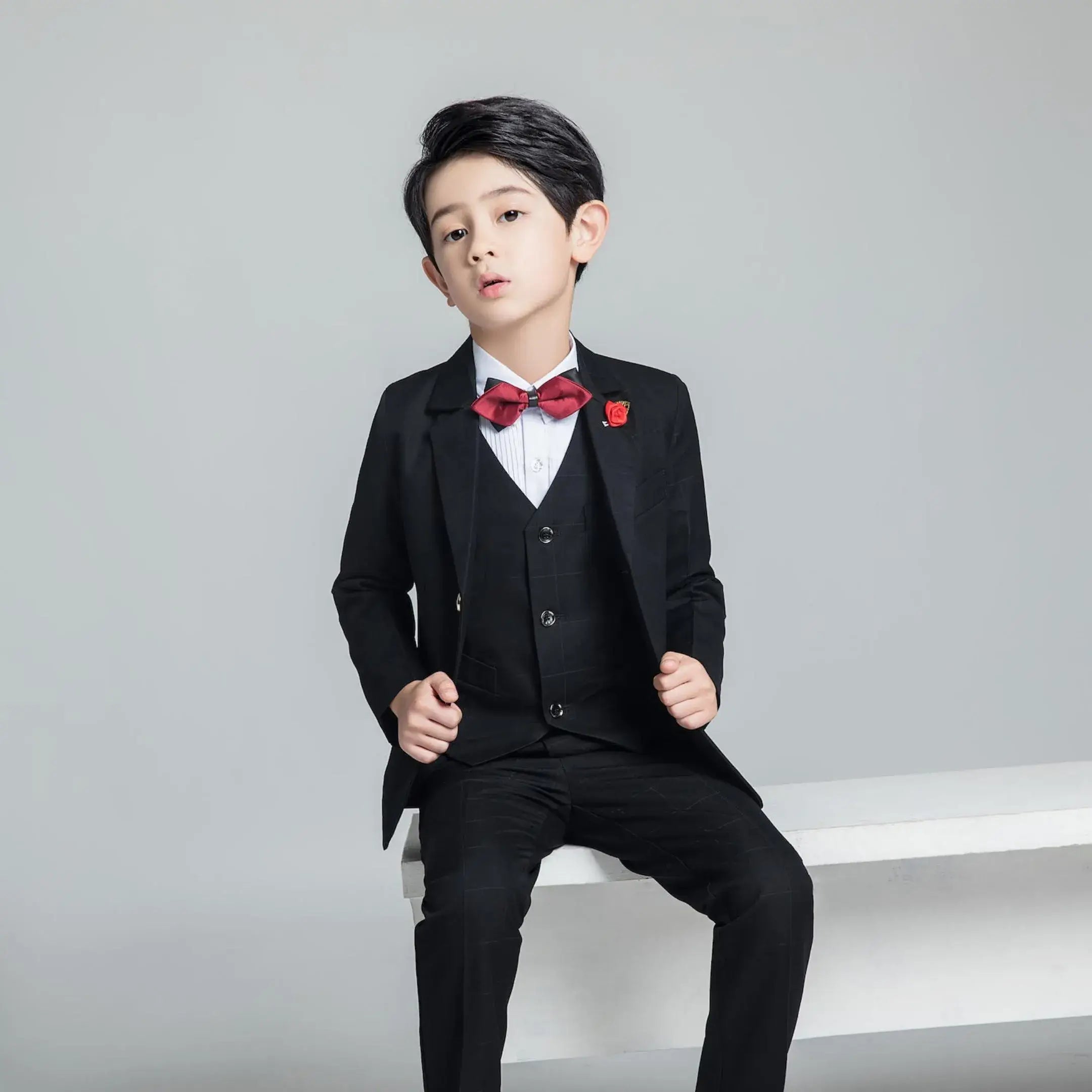 Boy sitting in black plaid 3-piece suit with red bow tie and boutonniere for weddings or formal events