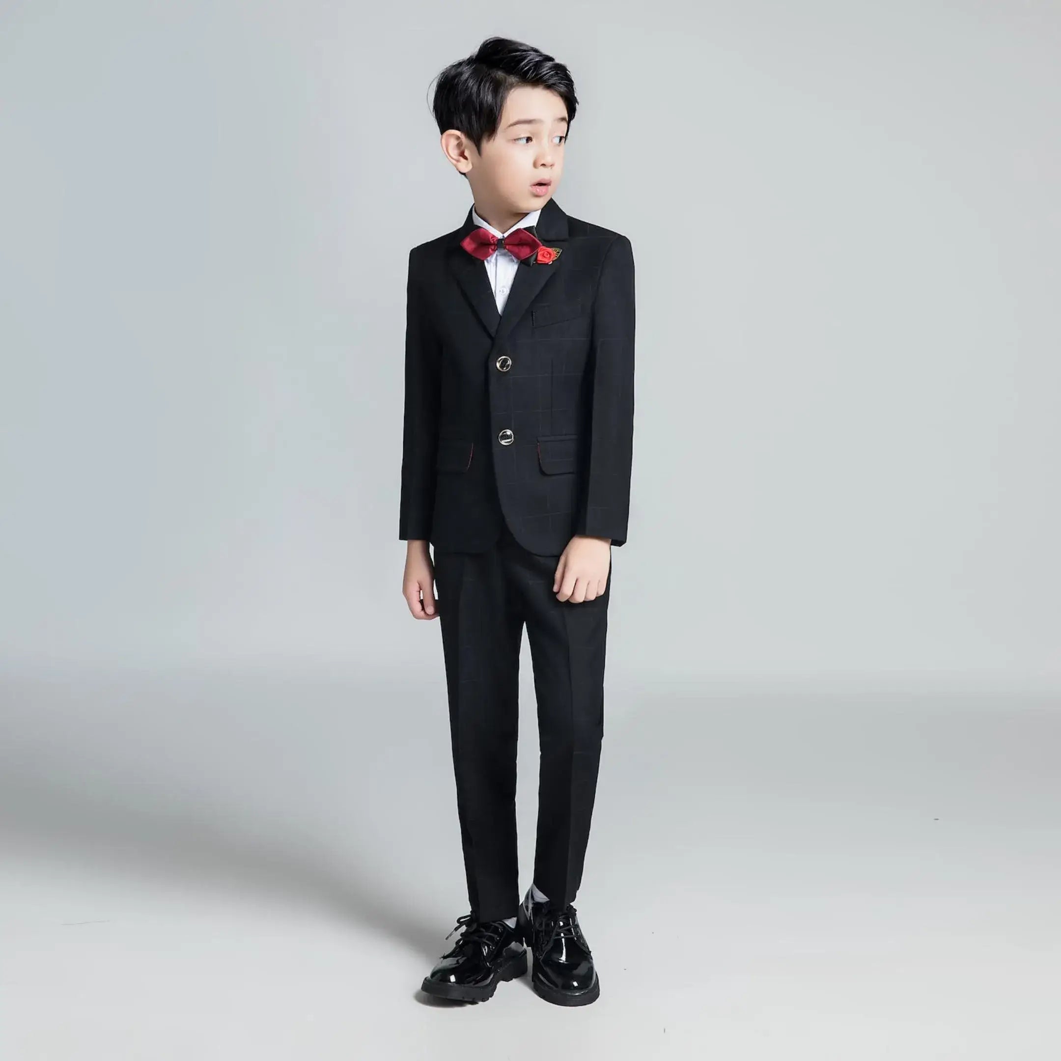 Boy in black plaid tuxedo with red bow tie and flower brooch posing for formal occasion