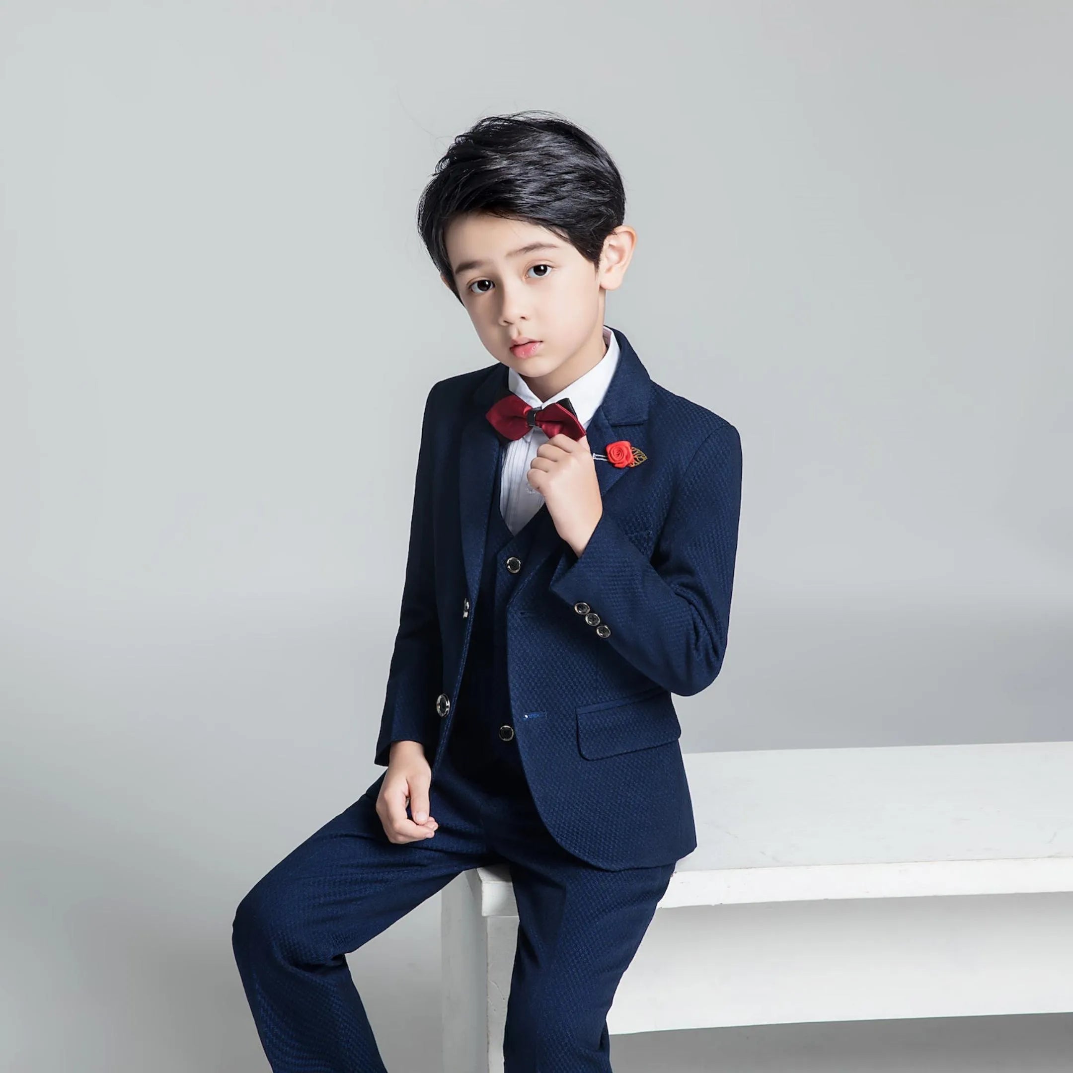 Boy in navy textured 3-piece suit adjusting red bow tie, seated stylishly in formal wedding outfit