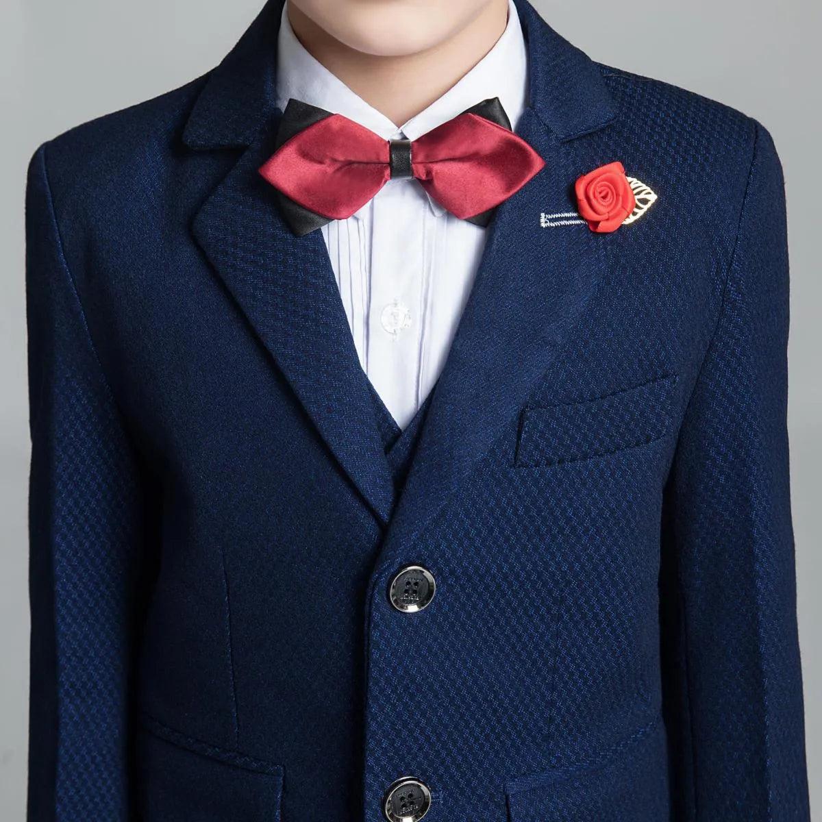 Close-up of navy textured boys' suit with red bow tie and rose pin, perfect for formal or wedding occasions