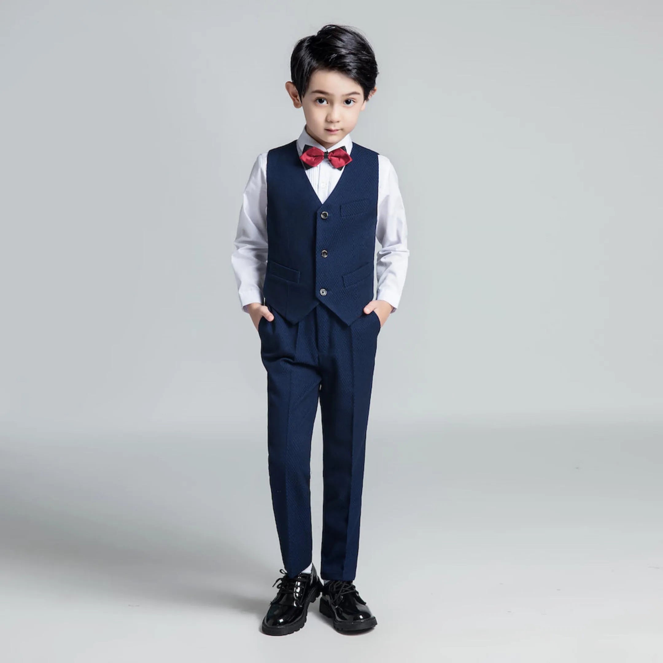 Young boy in navy vest and trousers with red bow tie, hands in pockets, dressed for wedding or formal event