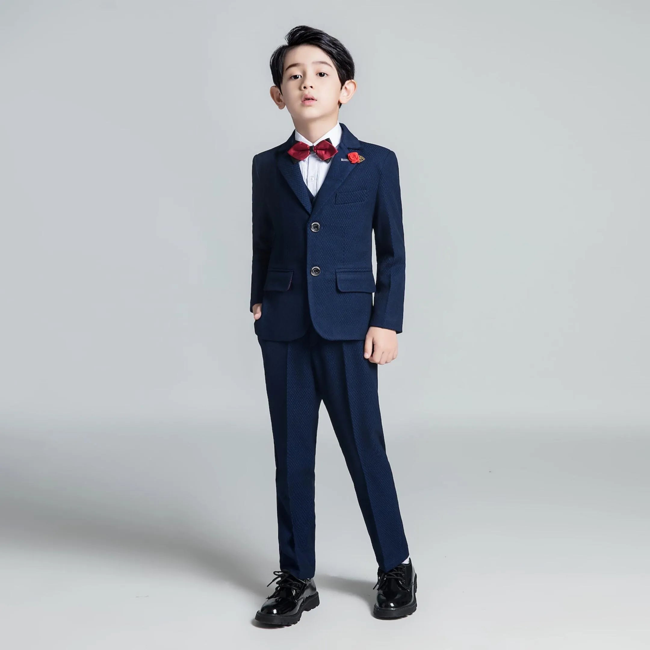 Boy posing in deep blue slim fit formal suit with bow tie and floral pin, styled for weddings or special events