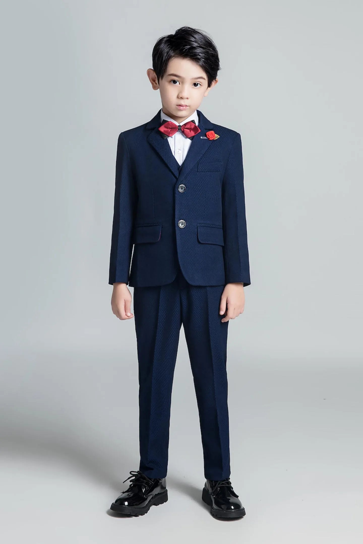 Full-body view of boy in slim fit navy blue textured suit with bow tie and floral lapel pin for formal wedding occasions