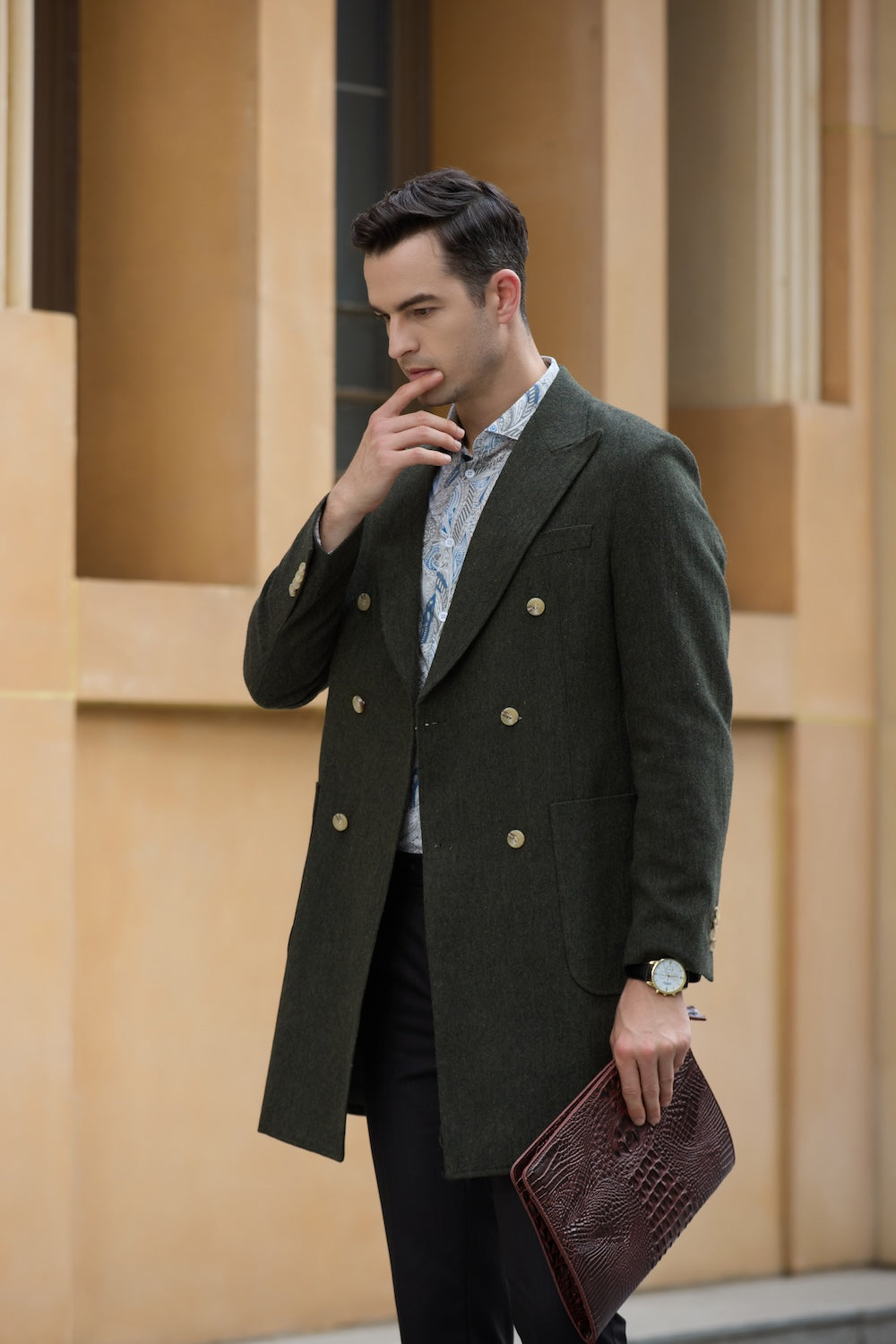 Man in long green overcoat holding textured clutch and thinking in city backdrop