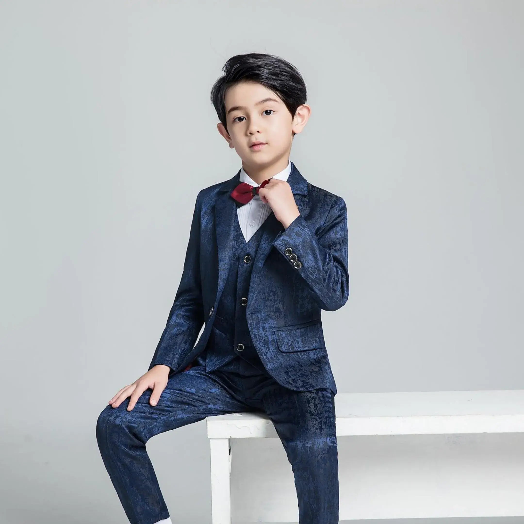 Boys' navy floral textured suit with red bow tie, sitting pose in elegant formal outfit for parties or weddings