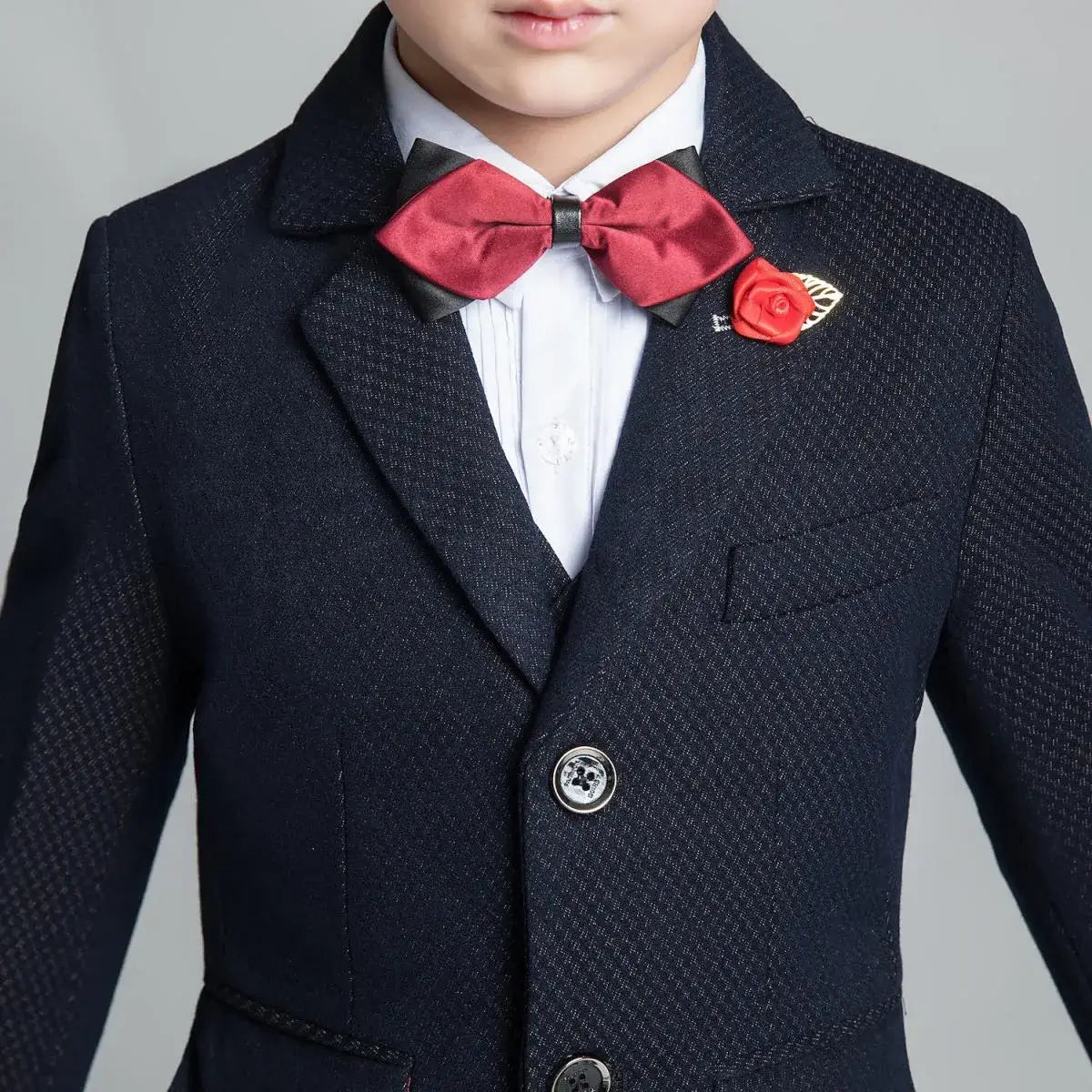 Close-up of navy boys' suit jacket with subtle textured pattern, red bow tie and rose pin for formal events and holiday wear
