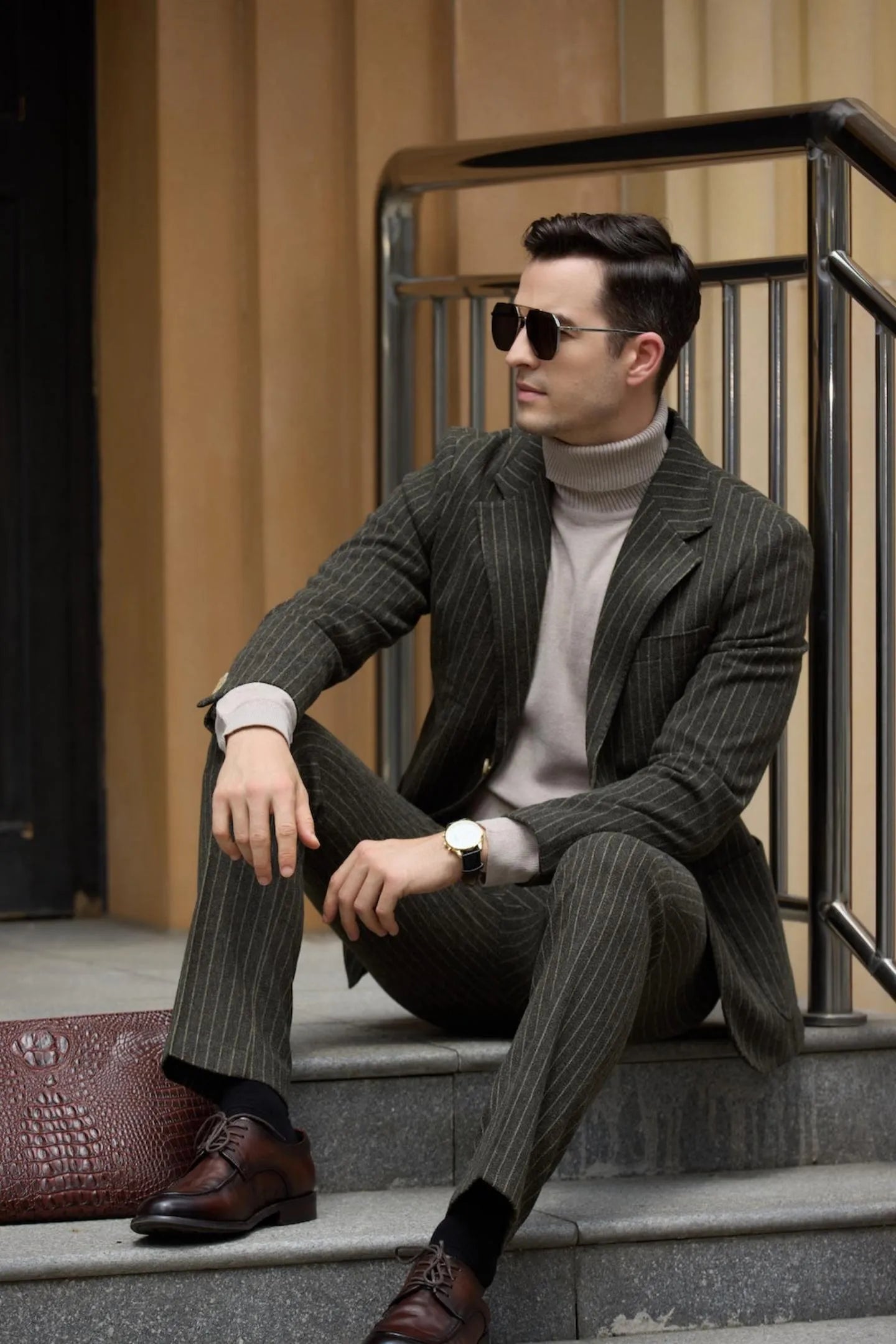 Man sitting on stairs, wearing a men's green herringbone pinstripe 2-piece suit, holding a clutch bag and exuding business elegance.