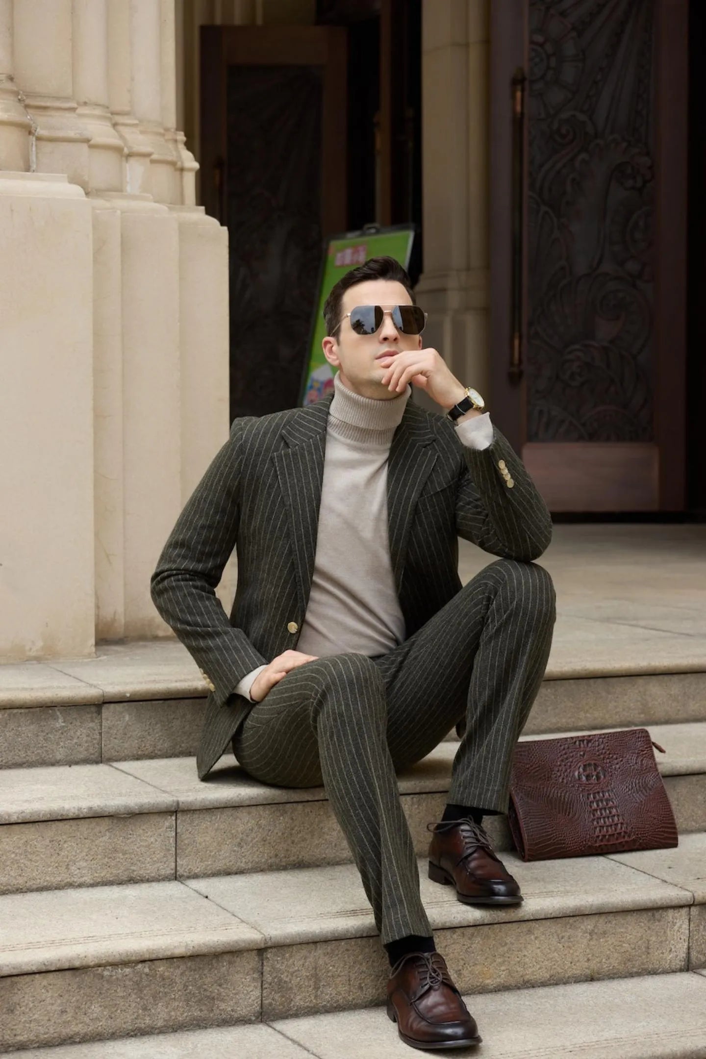 Man wearing a green herringbone pinstripe suit, with a beige turtleneck, sitting on stairs, looking confidently at the camera while holding sunglasses.