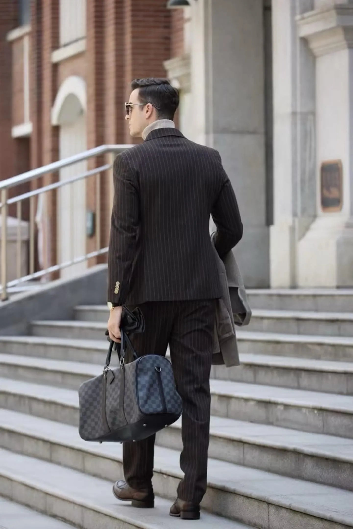Back view of men's coffee herringbone chalk stripe double-breasted suit – walking confidently with a business bag for a powerful professional look.